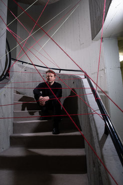 Eine Person sitzt auf einer Treppe in einem Betongebäude, umgeben von mehreren gespannten roten Fäden, die von Wand zu Wand verlaufen.