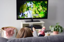 Frau hält Fernbedienung für den TV in der Hand. TV-Bild zeigt eine Nahaufnahme einer grünen Baumkrone